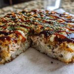 A slice cut from a round sushi bake, showing layers of rice, creamy filling, and topped with dark sauce and sesame seeds.