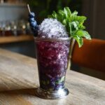 A vibrant Blueberry Mint Julep Cocktail served in a silver julep cup, topped with crushed ice, fresh mint, and skewered blueberries.