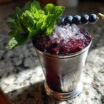 A refreshing Blueberry Mint Julep Cocktail served in a frosted silver julep cup, topped with crushed ice, fresh mint, and a skewer of blueberries.