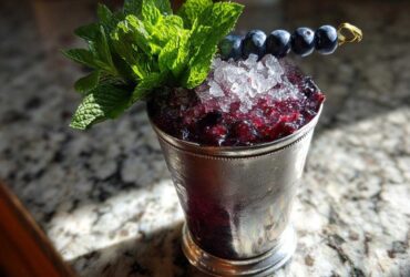 A refreshing Blueberry Mint Julep Cocktail served in a frosted silver julep cup, topped with crushed ice, fresh mint, and a skewer of blueberries.