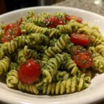 A close-up of spiral pasta tossed in vibrant green pesto, featuring halved cherry tomatoes in a white bowl, showcasing the Broccoli Pesto Pasta Salad.
