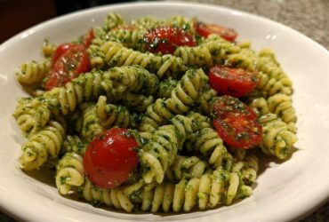 A close-up of spiral pasta tossed in vibrant green pesto, featuring halved cherry tomatoes in a white bowl, showcasing the Broccoli Pesto Pasta Salad.