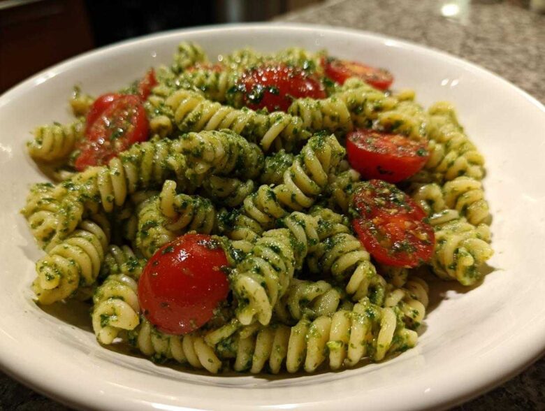 A close-up of spiral pasta tossed in vibrant green pesto, featuring halved cherry tomatoes in a white bowl, showcasing the Broccoli Pesto Pasta Salad.