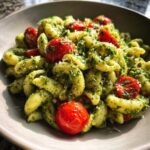 A close-up, appetizing shot of Broccoli Pesto Pasta Salad tossed with bright red cherry tomatoes in a grey bowl.