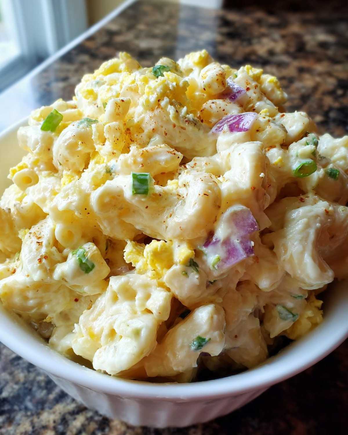 Close-up of creamy Classic Macaroni Salad in a white bowl, featuring elbow pasta, chopped egg, red onion, and green onion.