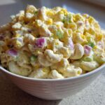 A close-up of a white bowl filled with creamy Classic Macaroni Salad (Potluck Style), featuring elbow pasta, celery, red onion, and chopped egg.