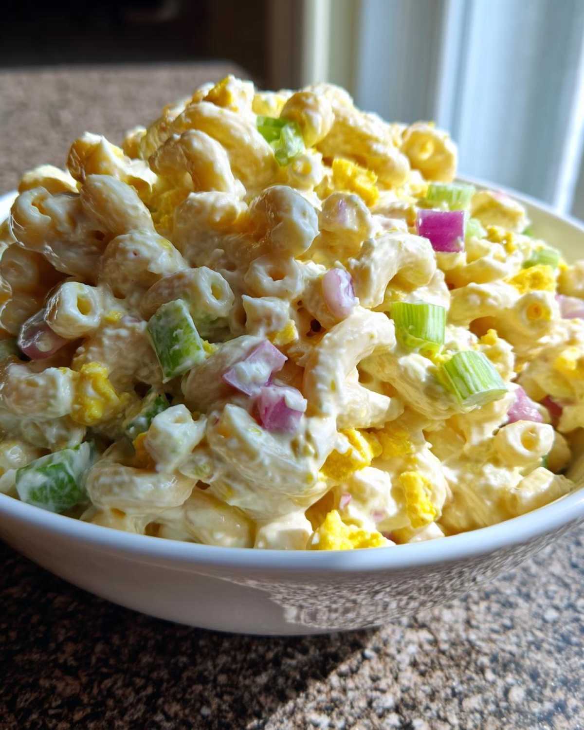 Close-up of a white bowl filled with creamy Classic Macaroni Salad, featuring elbow pasta, chopped celery, red onion, and hard-boiled egg.