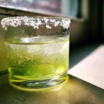 Close-up of a vibrant green Classic Margarita on the Rocks with a thick, frosty salt rim, sitting in sunlight.