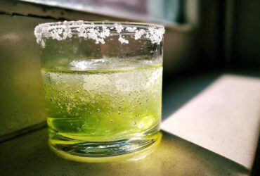 Close-up of a vibrant green Classic Margarita on the Rocks with a thick, frosty salt rim, sitting in sunlight.