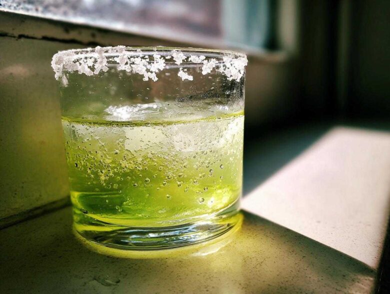 Close-up of a vibrant green Classic Margarita on the Rocks with a thick, frosty salt rim, sitting in sunlight.
