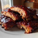 A stack of tender Classic Slow Cooker BBQ Ribs glazed with thick, shiny sauce resting on a white plate.