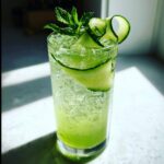 A tall glass filled with a vibrant green Cool Cucumber Mint Spritzer, garnished with fresh mint and cucumber ribbons.
