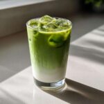A tall glass of vibrant green Creamy Iced Matcha Latte layered over white milk and filled with ice cubes.