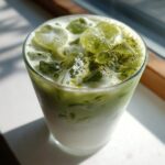 Close-up of a tall glass filled with a layered Creamy Iced Matcha Latte, topped with ice cubes and matcha powder.