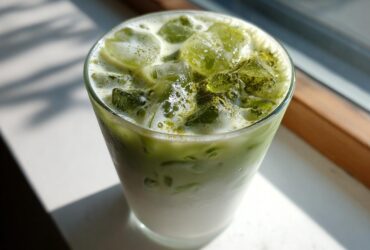 Close-up of a tall glass filled with a layered Creamy Iced Matcha Latte, topped with ice cubes and matcha powder.