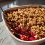 Close-up of a spoonful being taken from a white baking dish filled with bubbling Easy Cherry Crisp, showing the oat topping and bright red fruit.