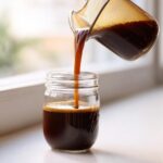 Pouring rich, dark Easy Homemade Cold Brew Coffee Concentrate from a pitcher into a glass mason jar.
