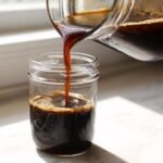 Dark, rich Easy Homemade Cold Brew Coffee Concentrate being poured from a glass container into a mason jar.