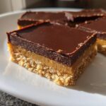 A close-up of a square Easy No-Bake Peanut Butter Chocolate Bars showing three distinct layers: crust, peanut butter filling, and chocolate topping.