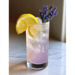 A tall glass filled with ice and pinkish-purple Fragrant Lavender Lemonade, garnished with lemon slices and fresh lavender sprigs.