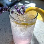 Close-up of a tall glass filled with ice and Fragrant Lavender Lemonade, garnished with fresh lavender sprigs and a lemon slice.