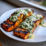 Two perfectly cooked Grilled Salmon Fillets topped generously with creamy lemon-dill sauce on a white plate.