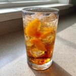 A tall glass filled with Homemade Fresh Peach Iced Tea, ice cubes, and slices of fresh peach, sitting in sunlight.