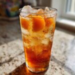 A tall, sweating glass filled with Homemade Fresh Peach Iced Tea, ice cubes, and peach slices.