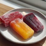 Three vibrant Homemade Fruit Popsicles (red, orange, and dark purple) resting on a speckled white plate in bright sunlight.