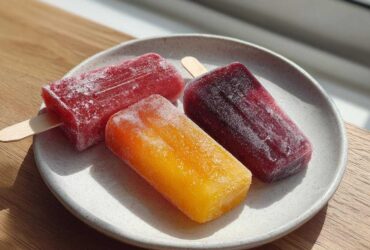 Three vibrant Homemade Fruit Popsicles (red, orange, and dark purple) resting on a speckled white plate in bright sunlight.