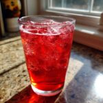 A tall glass filled with vibrant red Homemade Sparkling Raspberry Soda and ice cubes, illuminated by sunlight.