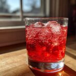 Close-up of a glass filled with bright red Homemade Sparkling Raspberry Soda and ice cubes, showing intense carbonation.