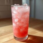A tall glass filled with bubbly, pink Hydrating Watermelon Agua Fresca and ice cubes, sitting on a wooden counter.