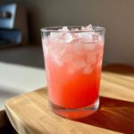 Close-up of a tall glass filled with bright pink Hydrating Watermelon Agua Fresca and crushed ice, sitting on a wooden board.