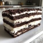 A thick slice of Layered Oreo Icebox Cake showing alternating dark chocolate cookie layers and white cream filling, topped with crumbs.