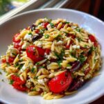 A close-up, vibrant bowl of Mediterranean Orzo Salad featuring pasta, bright red cherry tomatoes, feta cheese, and olives.