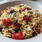A close-up view of a bowl filled with Mediterranean Orzo Salad featuring pasta, roasted cherry tomatoes, crumbled feta cheese, and herbs.