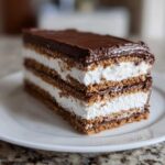 Close-up of a layered slice of No-Bake Chocolate Eclair Cake Dessert with graham crackers, white cream, and chocolate frosting.
