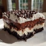 A decadent slice of No-Bake Chocolate Lasagna Dessert showing four distinct layers topped with whipped cream and mini chocolate chips.