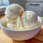 Three generous scoops of creamy No-Churn Vanilla Bean Ice Cream served in a white bowl on a wooden surface.