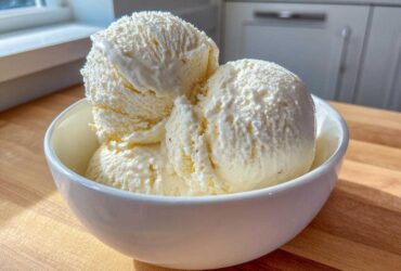 Three generous scoops of creamy No-Churn Vanilla Bean Ice Cream served in a white bowl on a wooden surface.