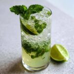 A tall glass filled with ice, mint leaves, and lime, representing The Perfect Classic Mojito Cocktail, with a lime wedge beside it.