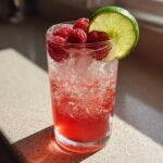 A tall glass filled with a fizzy, bright red Raspberry Lime Rickey, topped with fresh raspberries and a lime wheel.