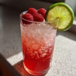 A tall glass filled with a vibrant red Raspberry Lime Rickey, crushed ice, fresh raspberries, and a lime wheel garnish.