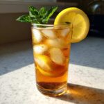 A tall glass filled with a Refreshing Arnold Palmer Drink, ice, and garnished with a lemon slice and fresh mint sprig.