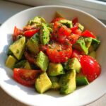 A close-up of a white bowl filled with a Refreshing Cucumber Tomato Avocado Salad, seasoned with herbs.