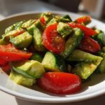 A close-up, sunlit bowl of Refreshing Cucumber Tomato Avocado Salad with vibrant red tomato wedges and green avocado chunks.