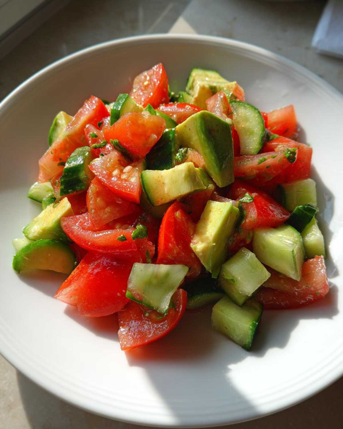 Close-up of a bowl filled with Refreshing Cucumber Tomato Avocado Salad, brightly lit by sunlight.