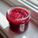Close-up of bright red Small Batch Quick Raspberry Jam glistening in a small glass jar sitting on a windowsill.