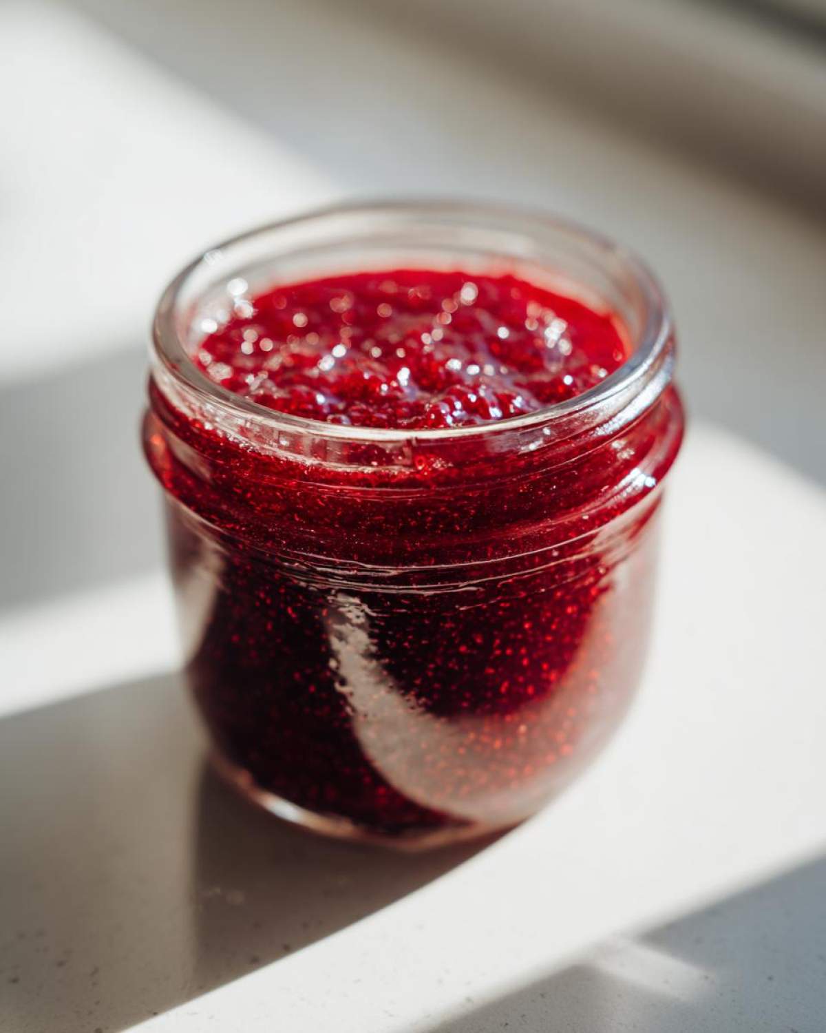 Close-up of a small glass jar filled with vibrant red Small Batch Quick Raspberry Jam, glistening in bright sunlight.